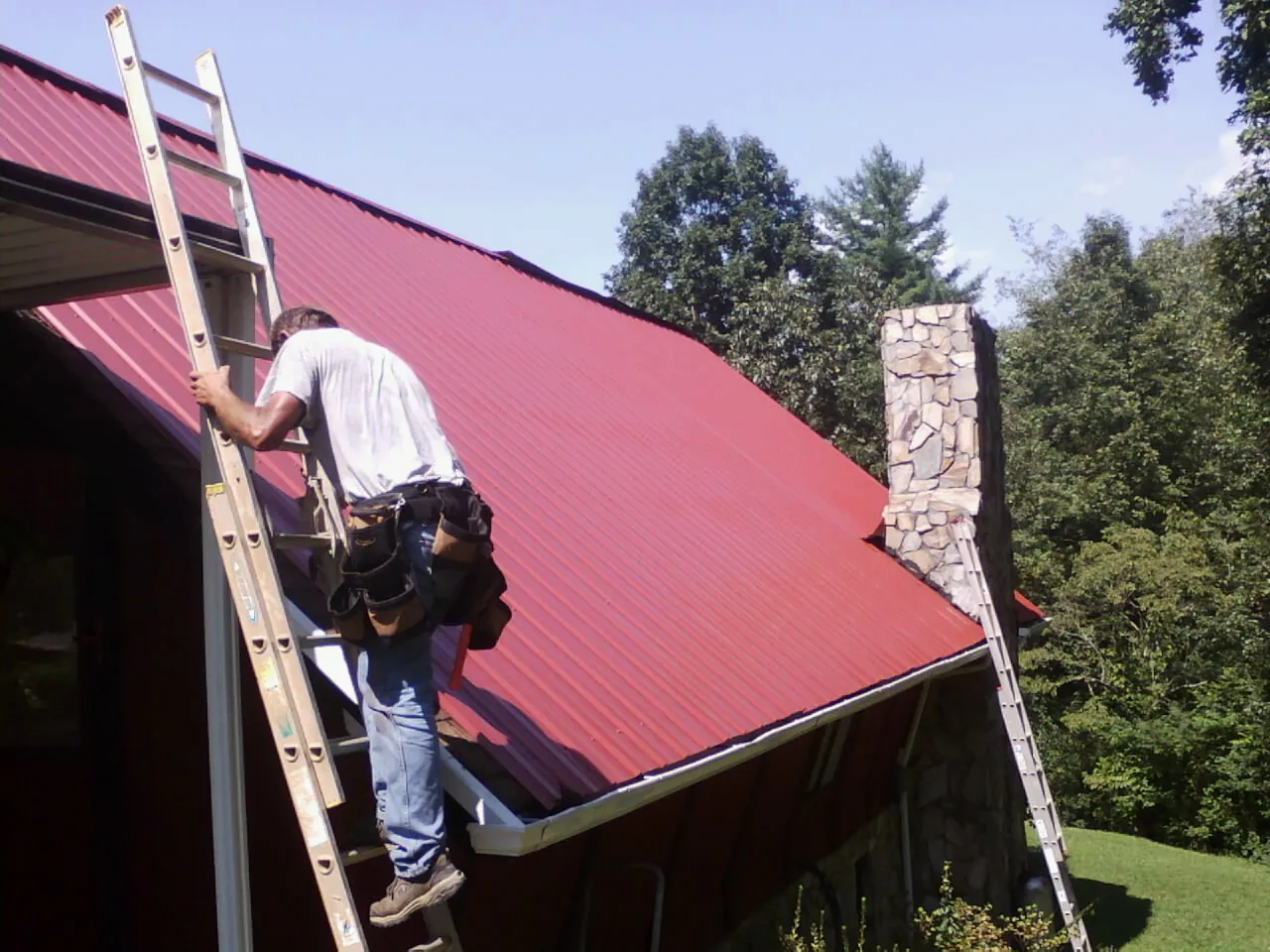 Roofer on a ladder beside a red metal roof and stone chimney — Soffit & Fascia Repair by Roofing Brothers in Aliquippa